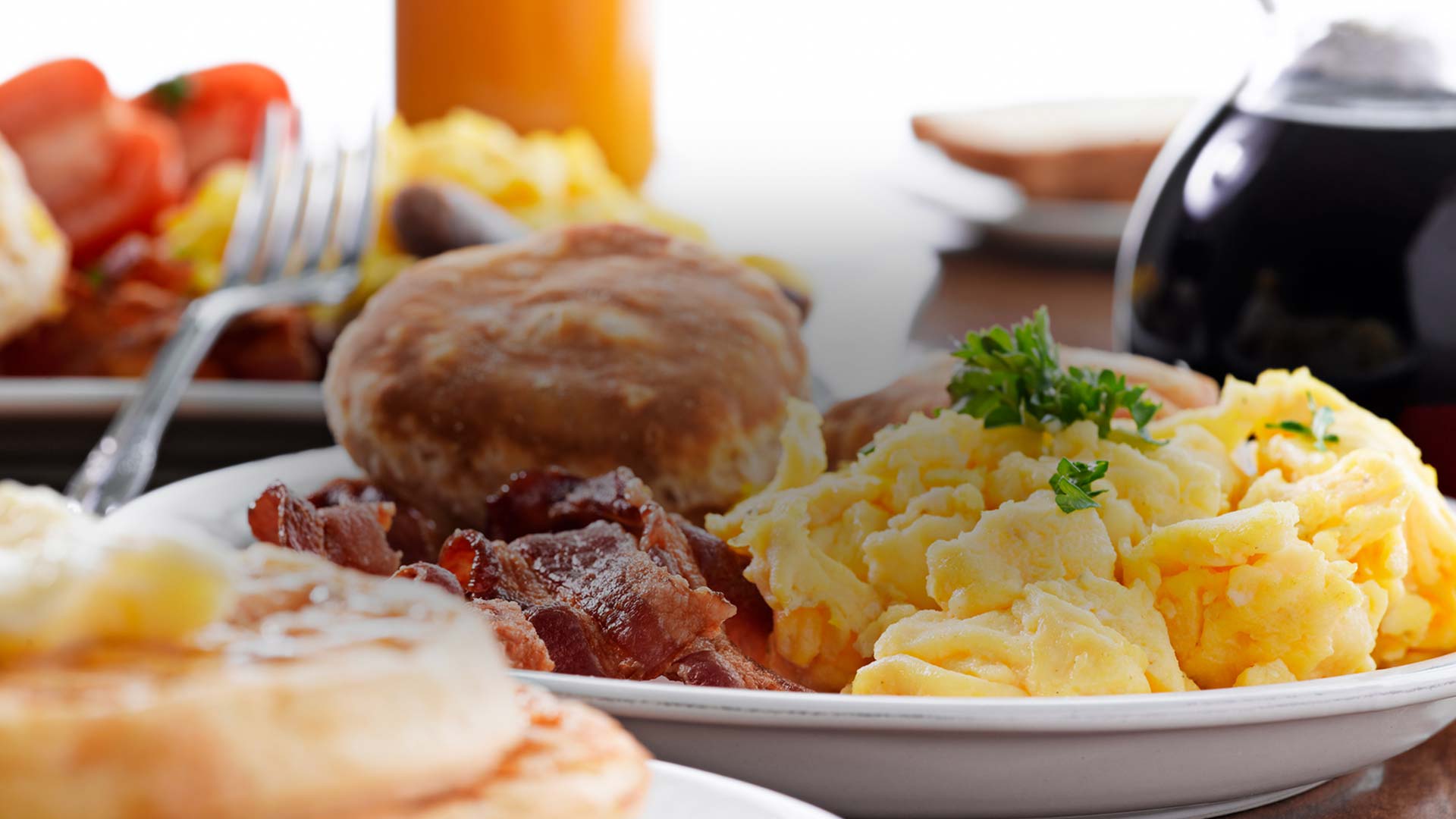 Scrambled eggs, bacon, and a biscuit on a plate, with pancakes and syrup in the background served at E.J.'s Diner in Wausau, WI.