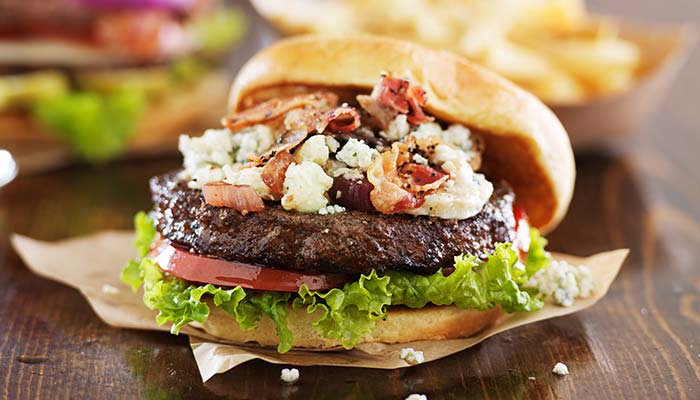 Burger with lettuce, tomato, bacon & blue cheese on a bun from E.J.'s Diner Menu; fries behind.