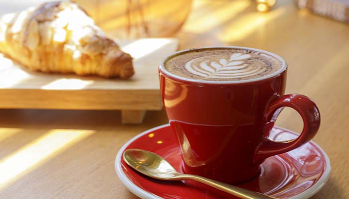 A red cup of latte and croissant from E.J.'s Diner Menu on a sunlit wooden table.